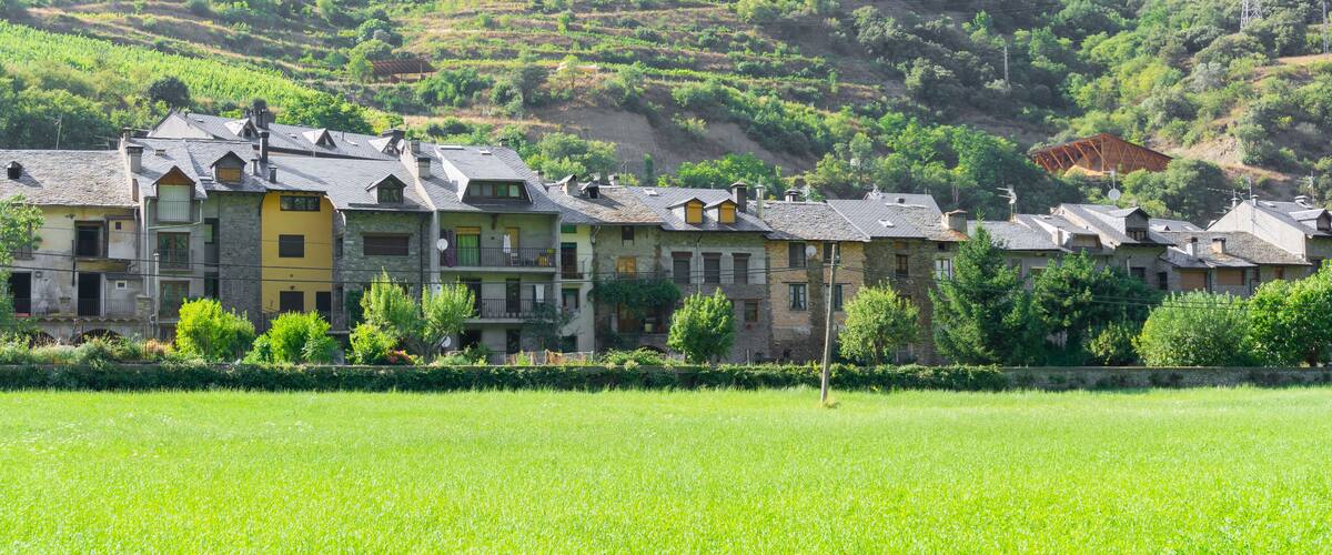 A row of traditional stone buildings with sloped roofs, set against a backdrop of lush green hills and a cultivated field.