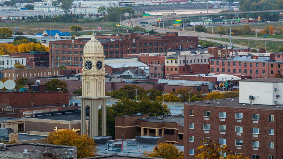 A cityscape with a prominent clock tower, a mix of red and white buildings, and a highway in the background.