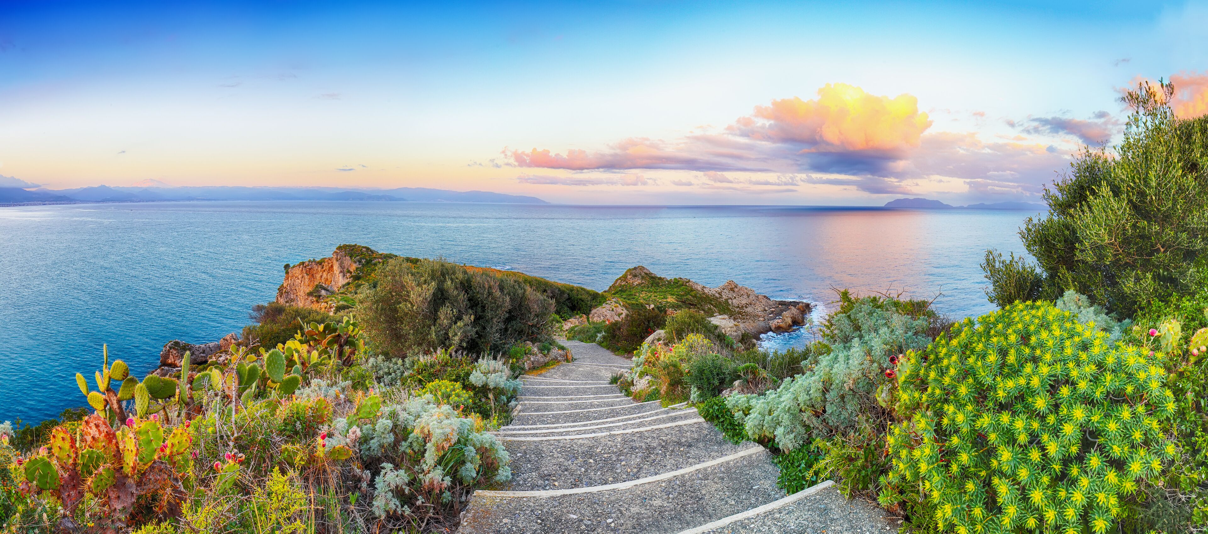 A coastal landscape with a stone staircase leading to a rocky outcrop, surrounded by lush greenery and overlooking the sea.