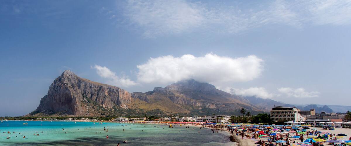 A beach with clear turquoise waters, a large rocky mountain, and a crowded beach area with umbrellas.