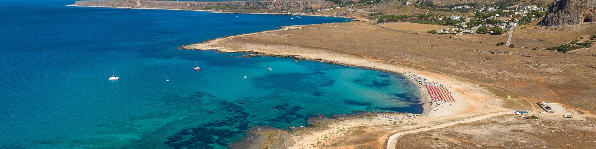 Paisagem costeira com águas azul-claras, uma praia arenosa e montanhas ao fundo.