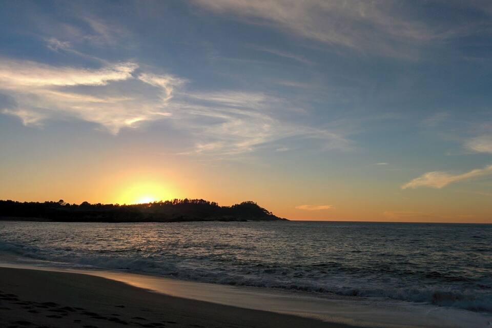 A sunset over a beach with waves and a distant tree-covered landmass.
