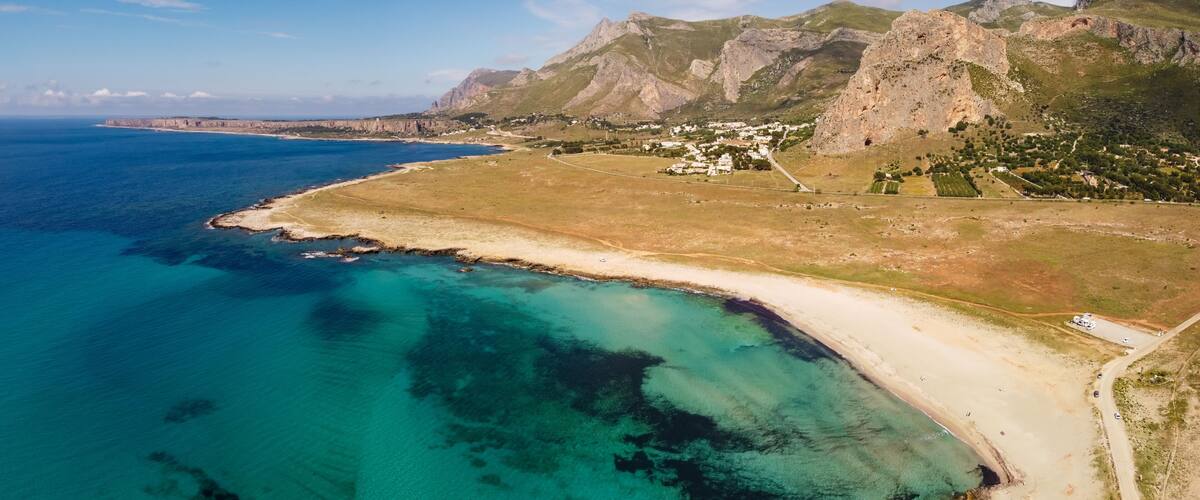 A coastal landscape with a clear blue sea, a sandy beach, and a mountainous backdrop.