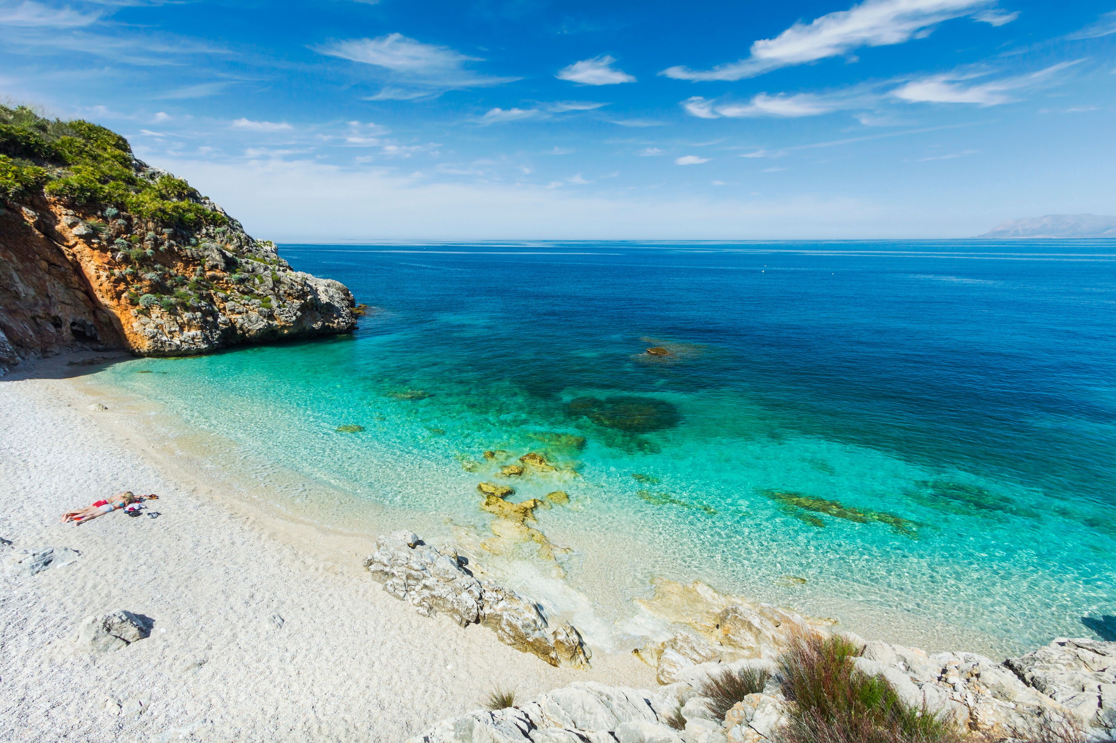 A clear turquoise sea, white sandy beach, rocky cliffs, and a clear sky.