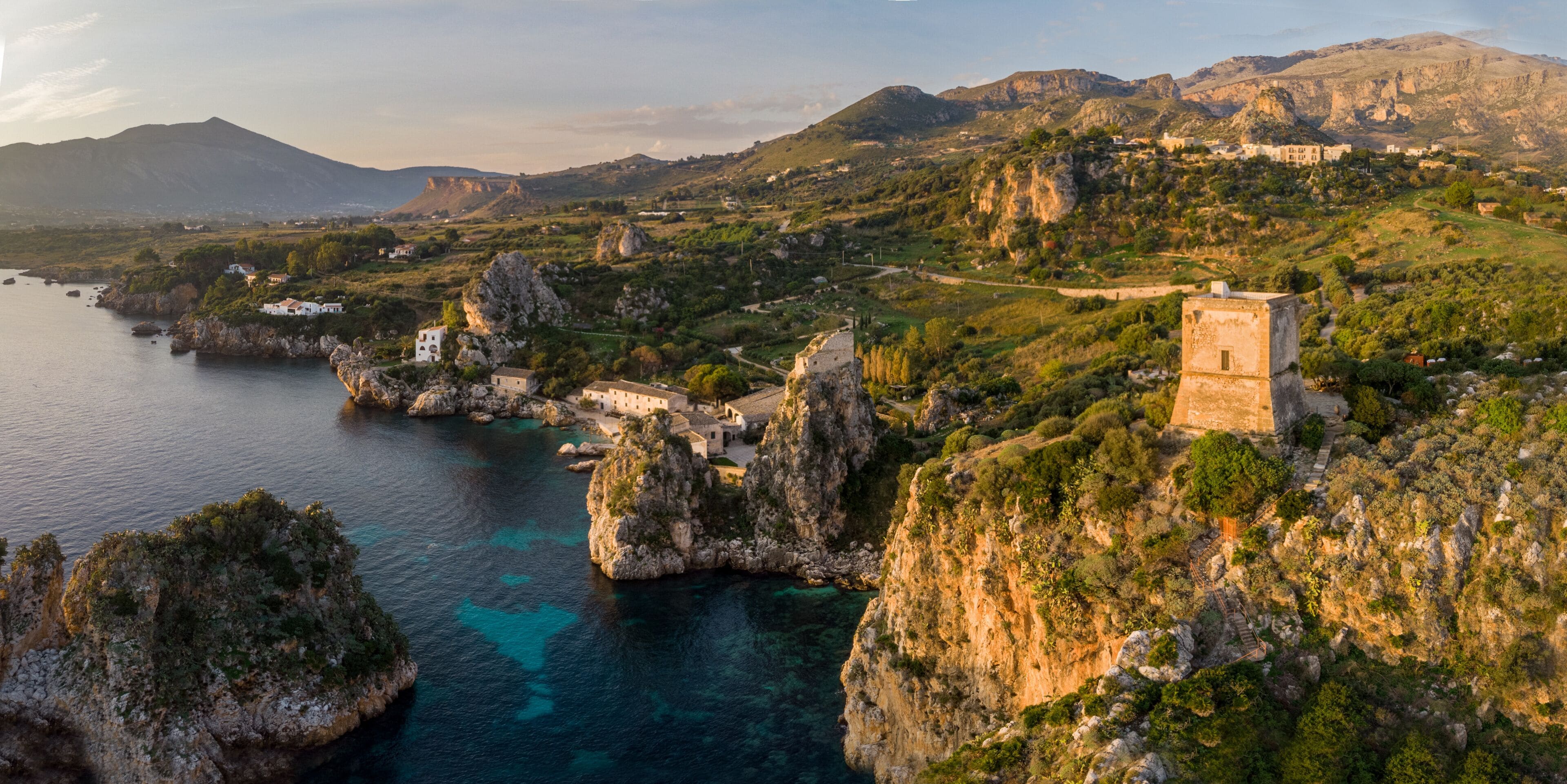 A coastal landscape with rocky cliffs, a clear blue sea, and a historic building on a hill.