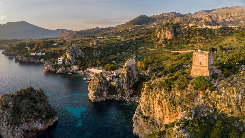 A coastal landscape with rocky cliffs, a clear blue sea, and a historic building on a hill.