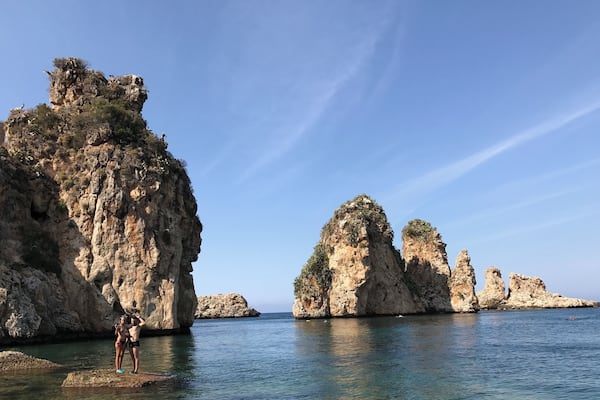 A rocky coastline with clear blue water and a couple standing on a rock.