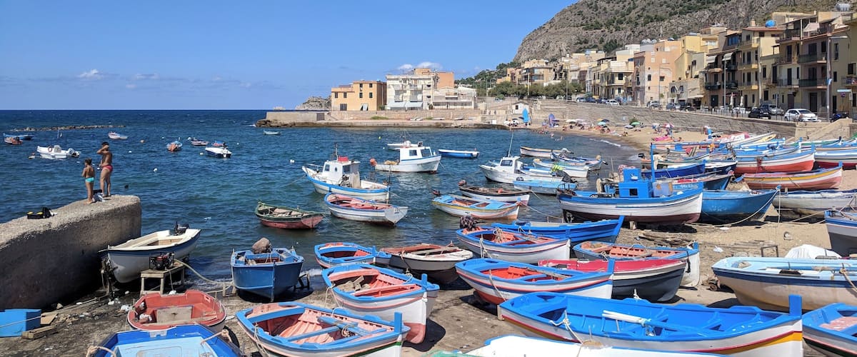 A coastal village with colorful boats docked along the shore, a rocky hillside, and traditional buildings.