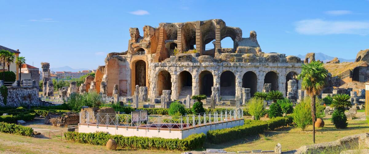 Ancient ruins with arches and columns, surrounded by greenery and modern buildings.