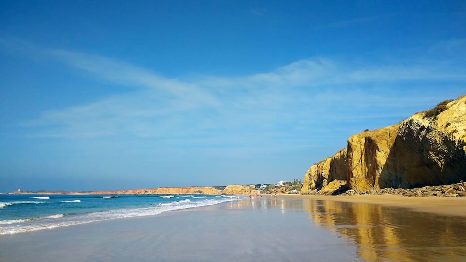 A beach with clear blue water, a sandy shore, and a rocky cliff.