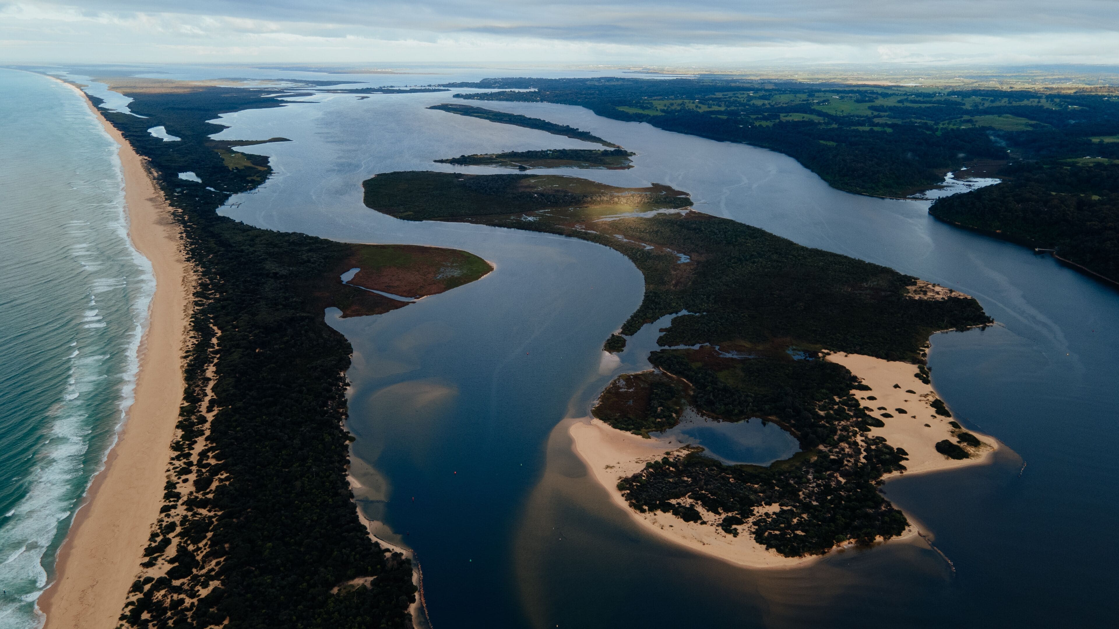 Un paisaje costero con una playa arenosa, una gran masa de agua y una zona boscosa.