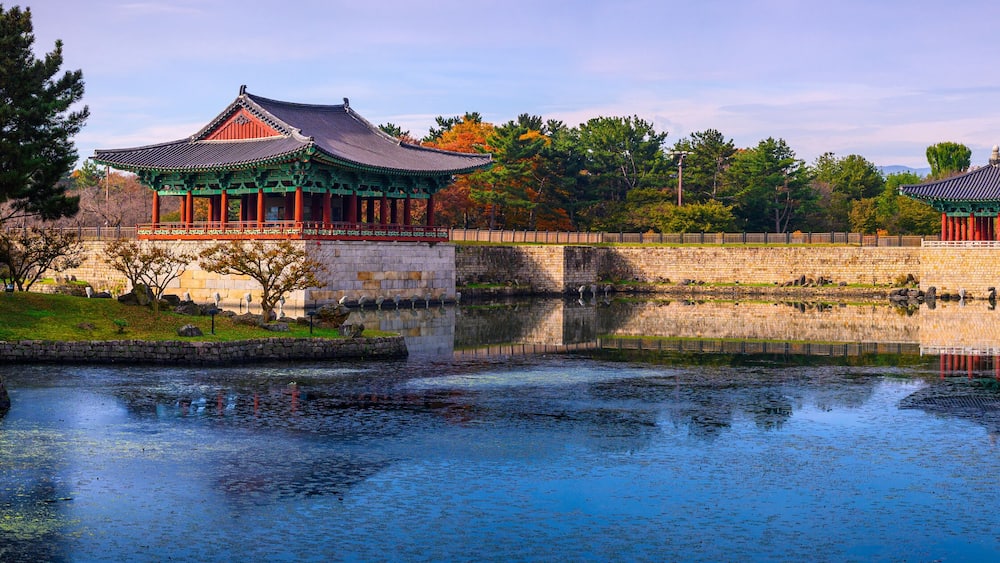 Traditional Korean architecture with a pond and autumn foliage.