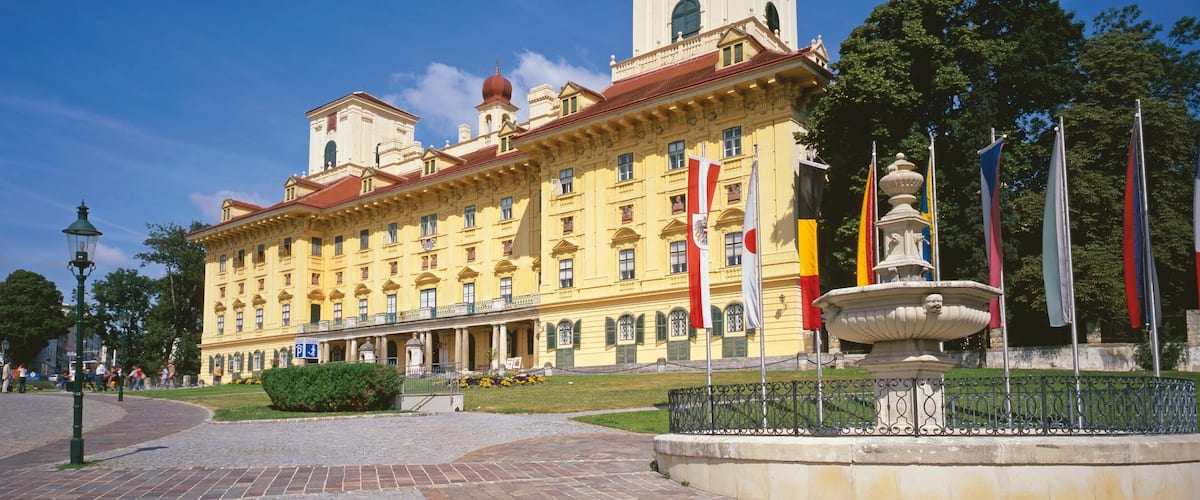 A large yellow building with a clock tower, surrounded by a paved area with a fountain and flags.