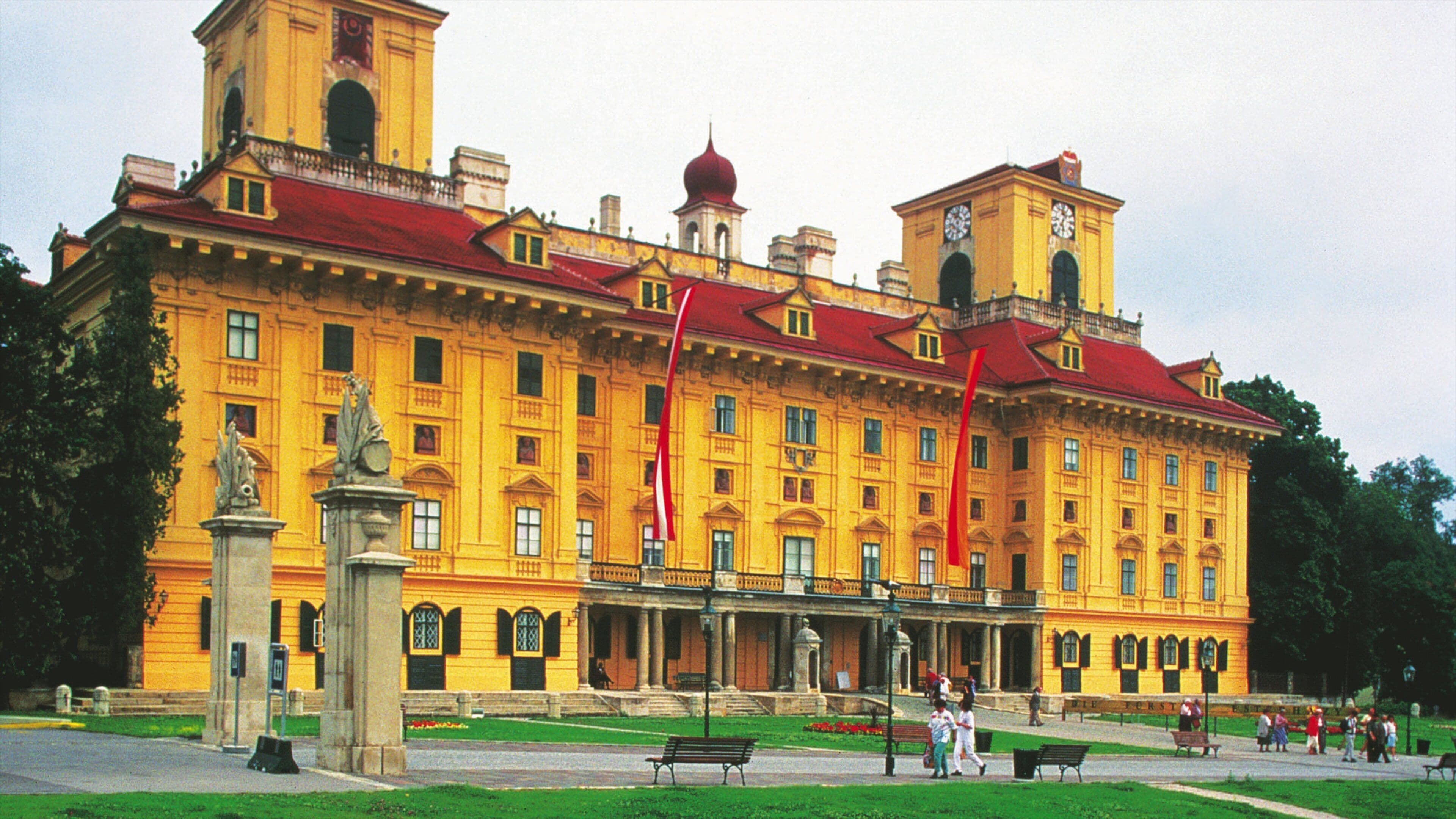 A large yellow building with a red roof, a clock tower, and a statue in front.
