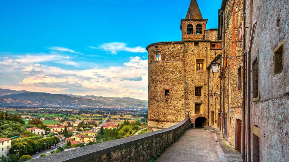 A historic stone tower with a conical roof overlooking a cobblestone pathway and a village below.