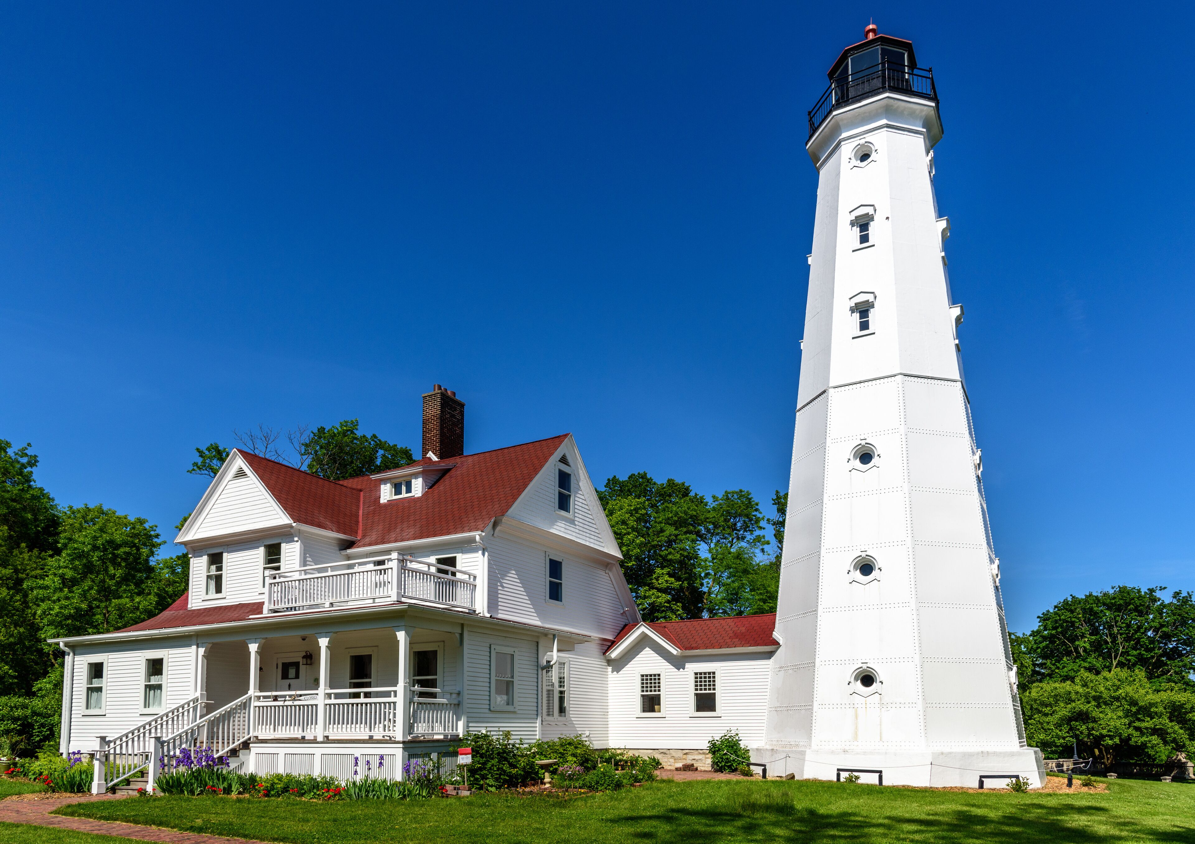 A white lighthouse with a red roof next to a white house with a porch.