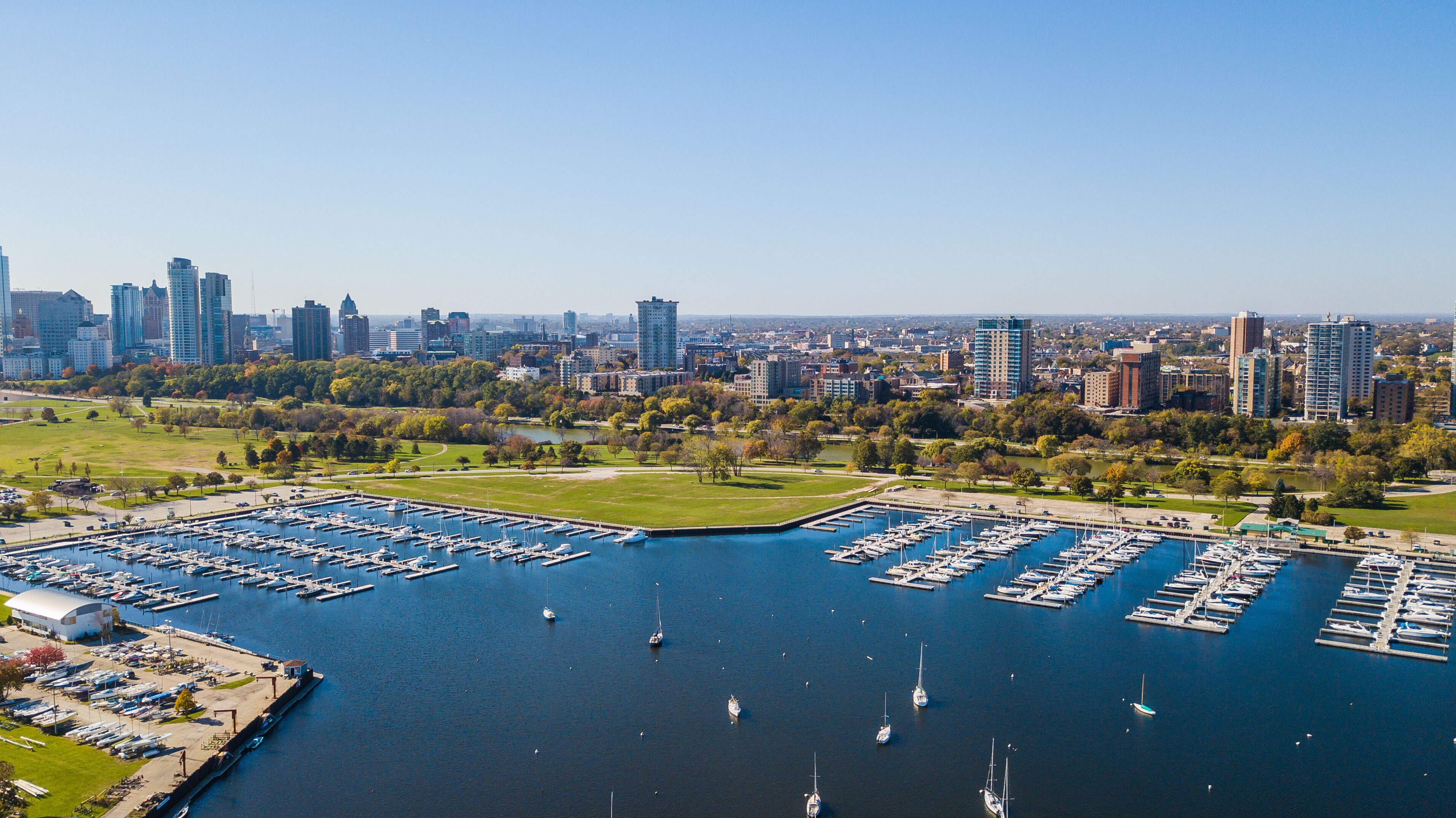 A marina with numerous boats docked, surrounded by a cityscape with high-rise buildings and a park with trees.