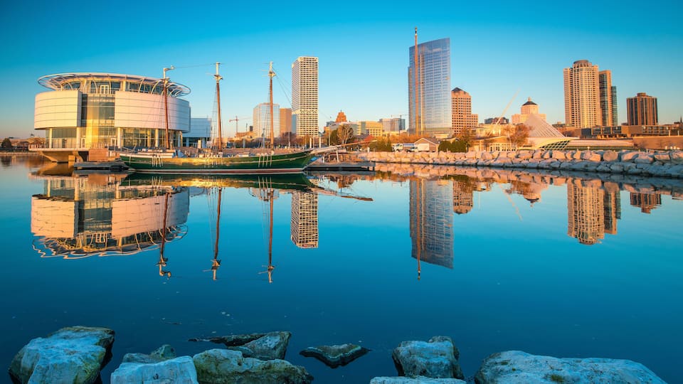 A waterfront cityscape with a modern building, a green boat, and tall skyscrapers reflecting in the water.