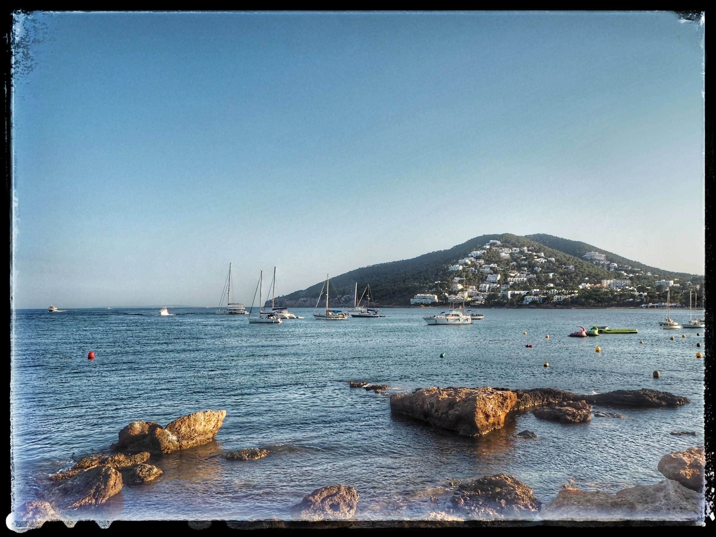 Escena costera con barcos, costa rocosa y una colina con edificios.