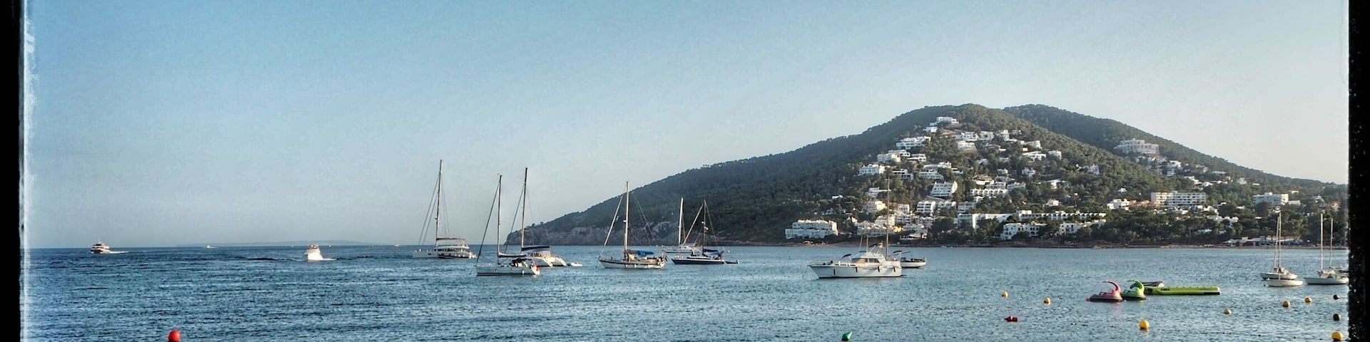 Escena costera con barcos, costa rocosa y una colina con edificios.