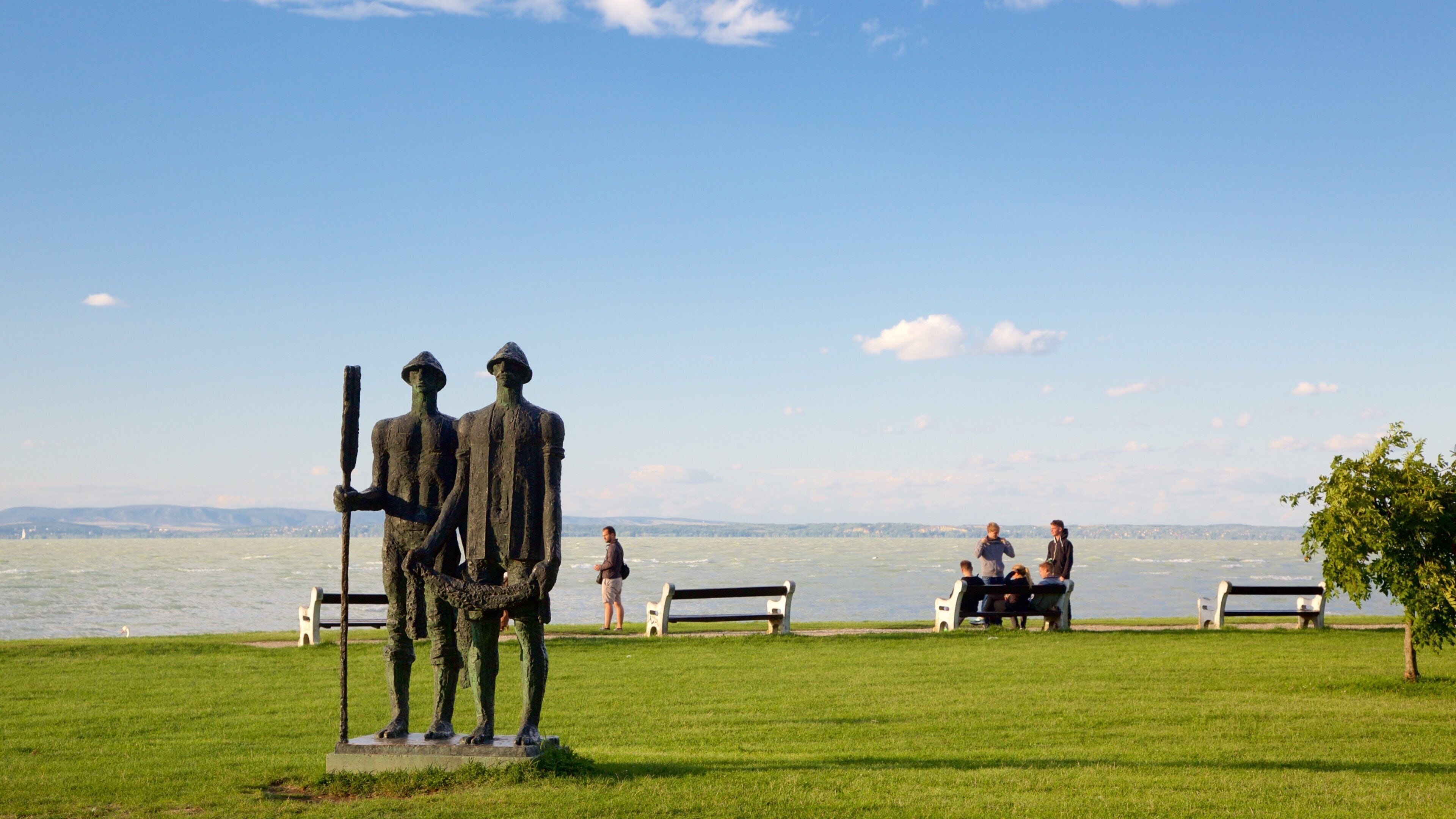 A statue of two figures on a grassy area by a lake with benches and people in the background.