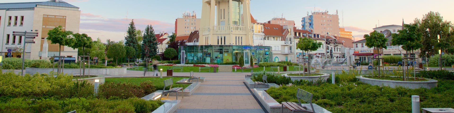 A tall tower with a glass facade in a city square surrounded by greenery and buildings.