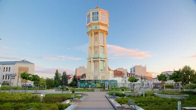 Uma torre alta com fachada de vidro em uma praça da cidade, cercada por vegetação e edifícios.