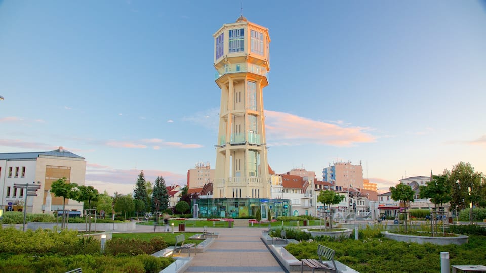 A tall tower with a glass facade in a city square surrounded by greenery and buildings.