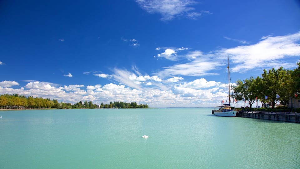 A calm sea with a sailboat docked at a pier, surrounded by trees and a clear blue sky.