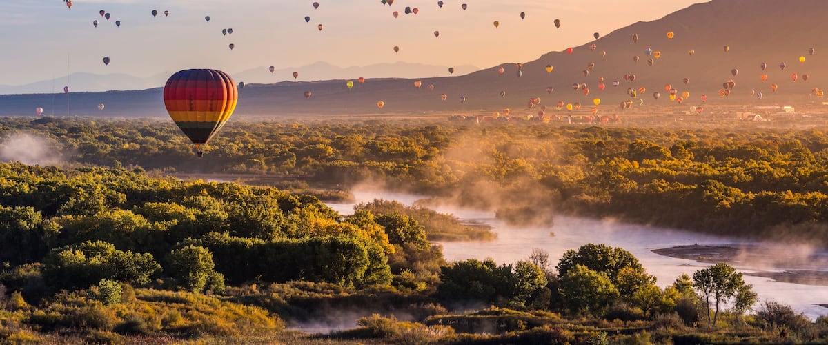 A hot air balloon festival with numerous balloons in the sky over a lush landscape with a river and mountains in the background.