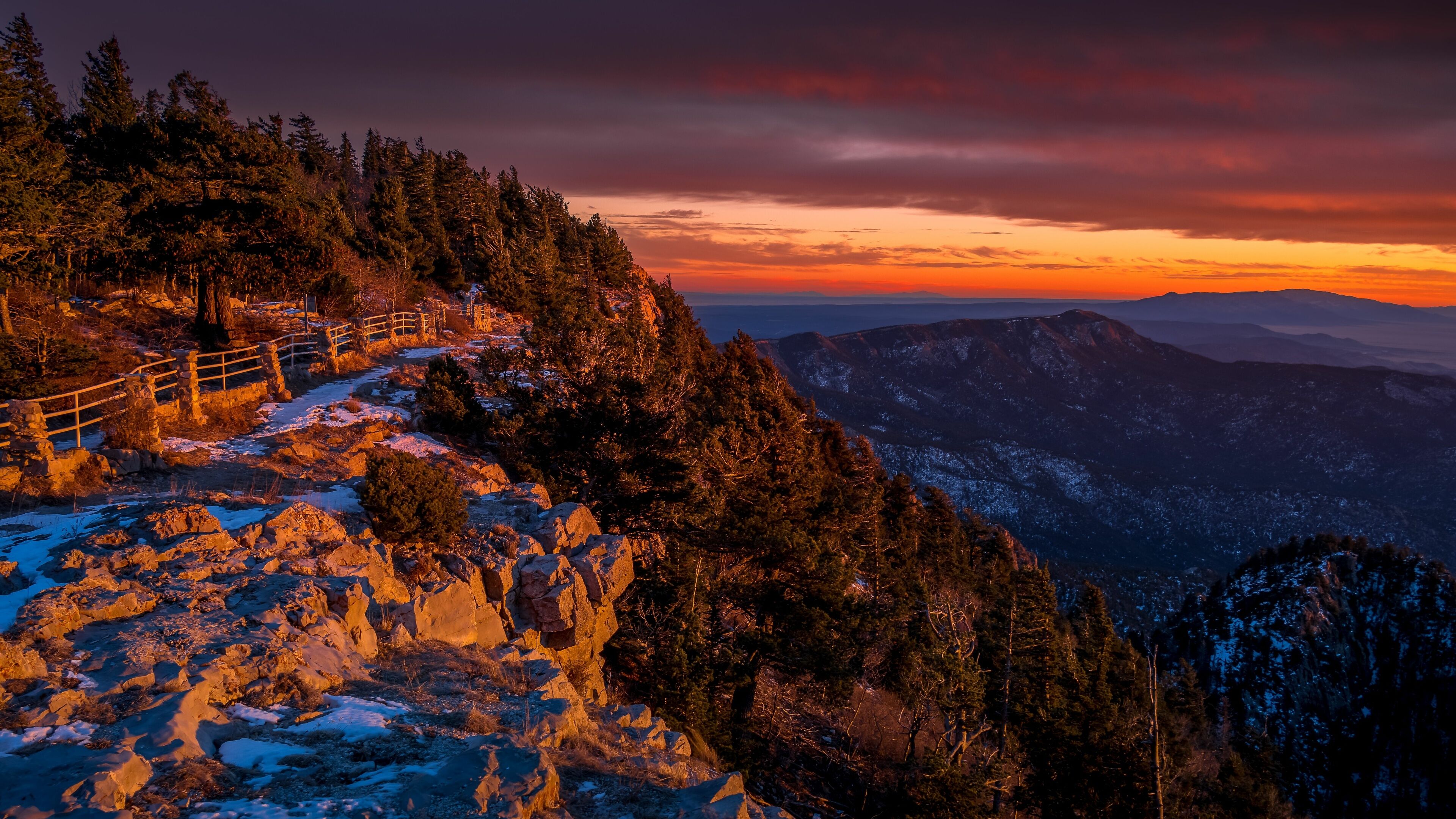 A snow-covered mountain path with a wooden fence, surrounded by dense forest and a sunset sky.