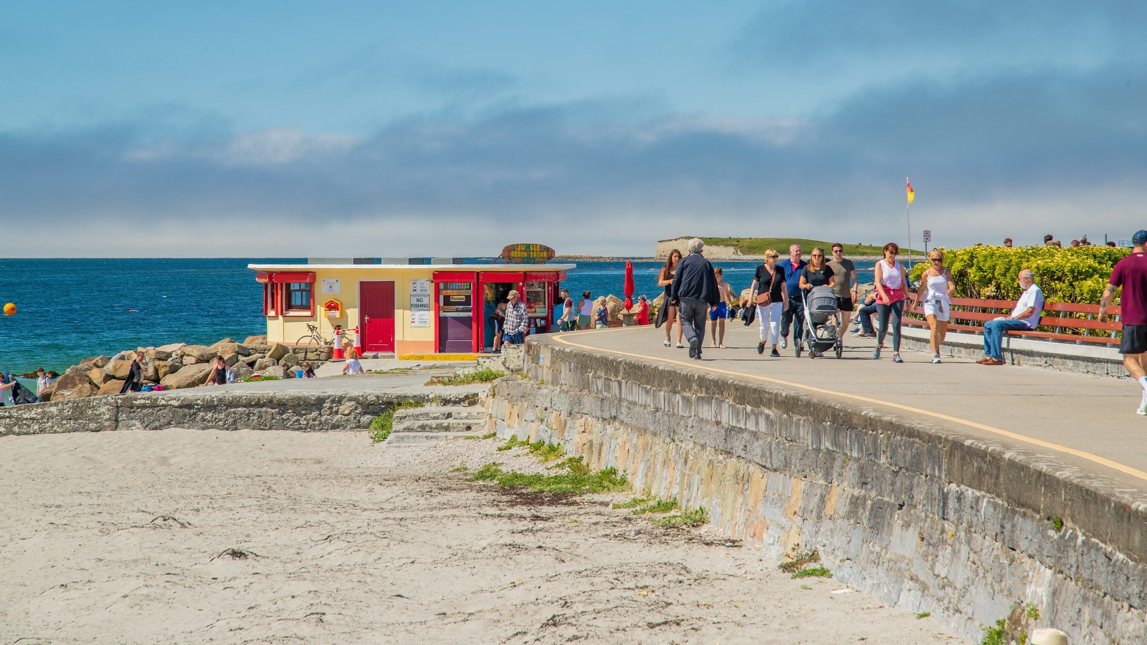 A coastal walkway with a lifeguard station, rocky shoreline, and people walking along the path.