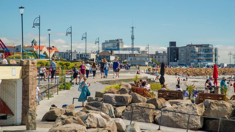 A coastal promenade with people walking, large rocks, and buildings in the background.