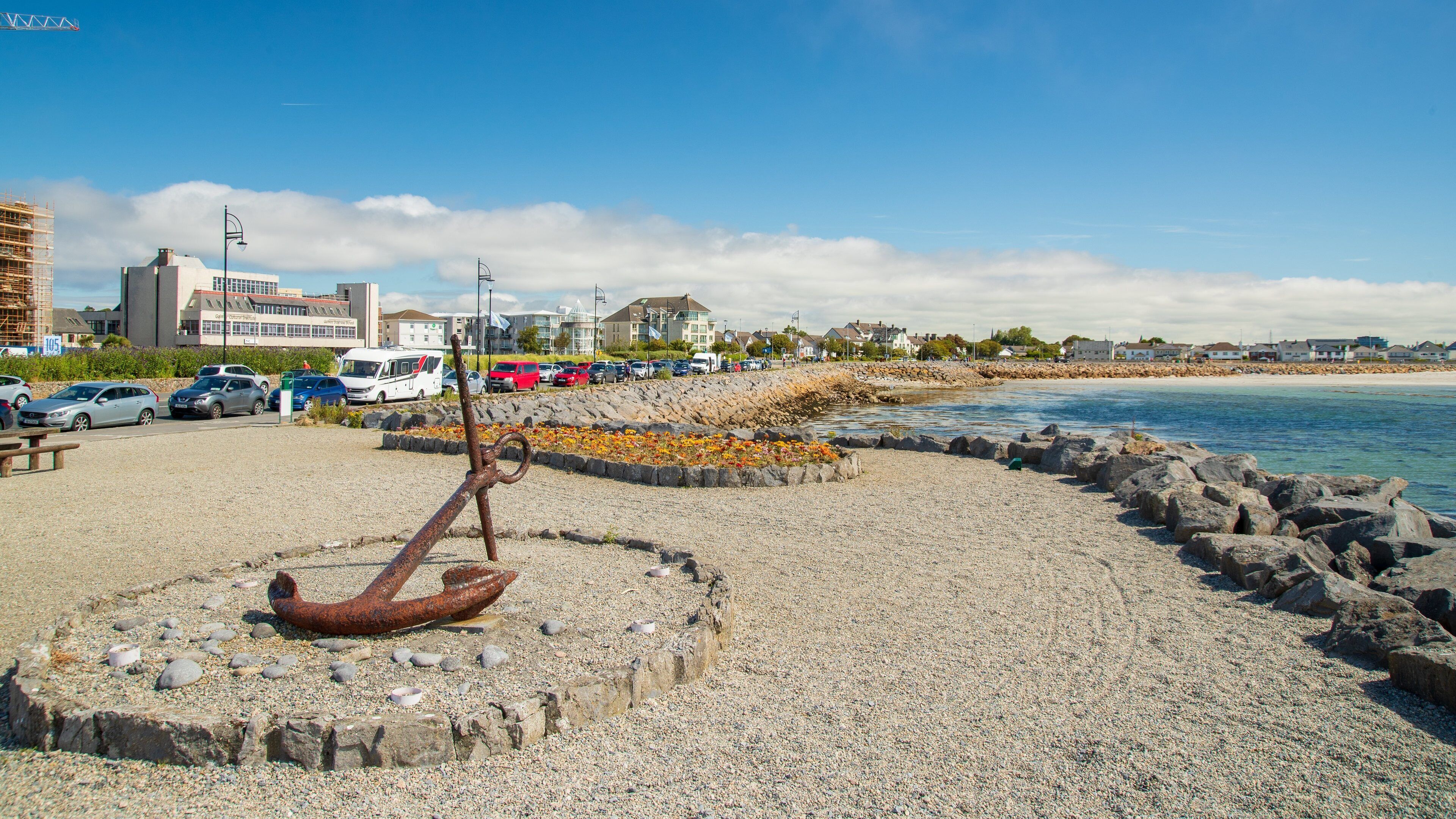 A pebbled beach with a rusty anchor sculpture, a rocky shoreline, and a row of buildings in the background.