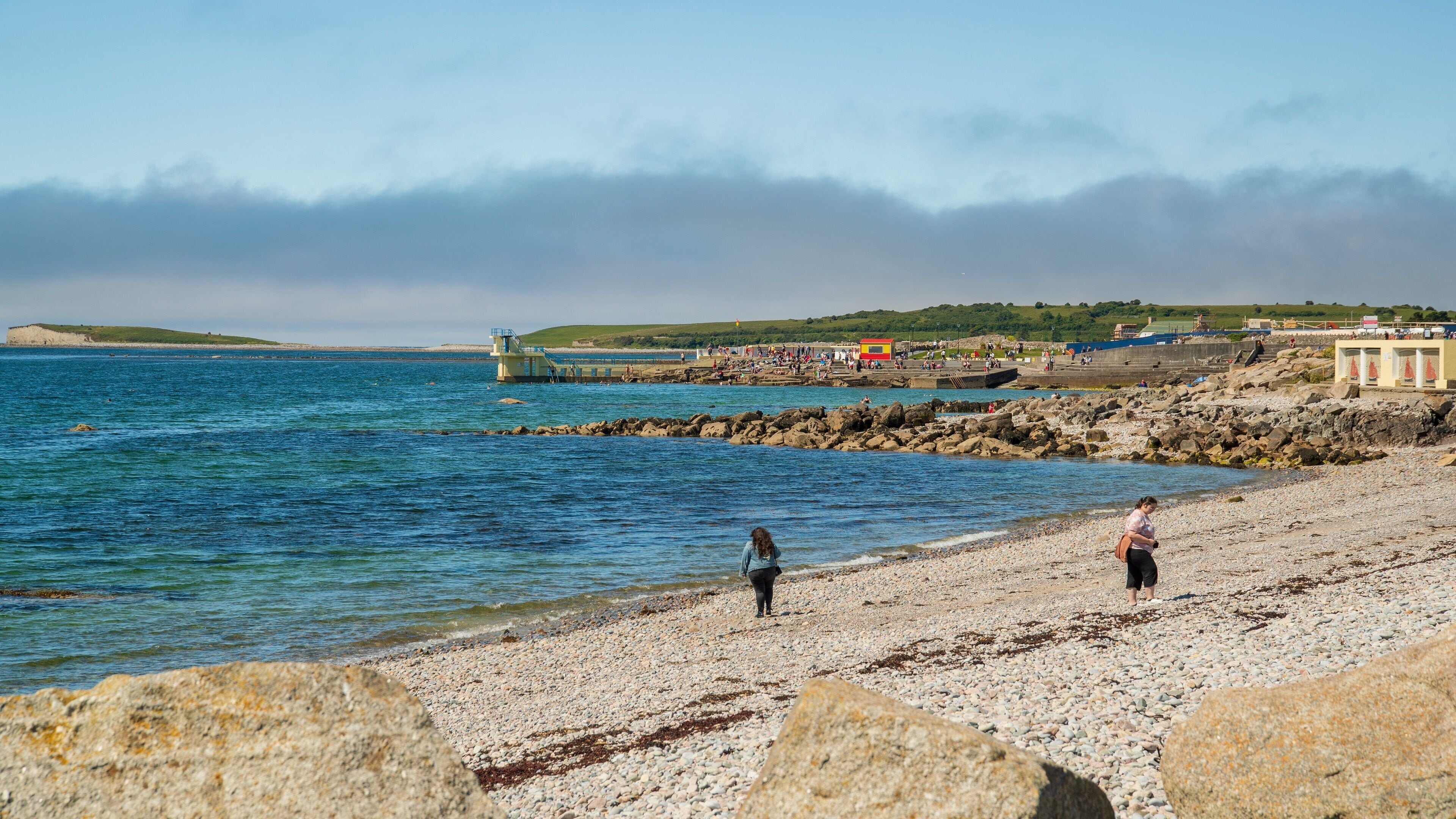 A pebbled beach with rocky outcrops, calm sea, and a distant hill.