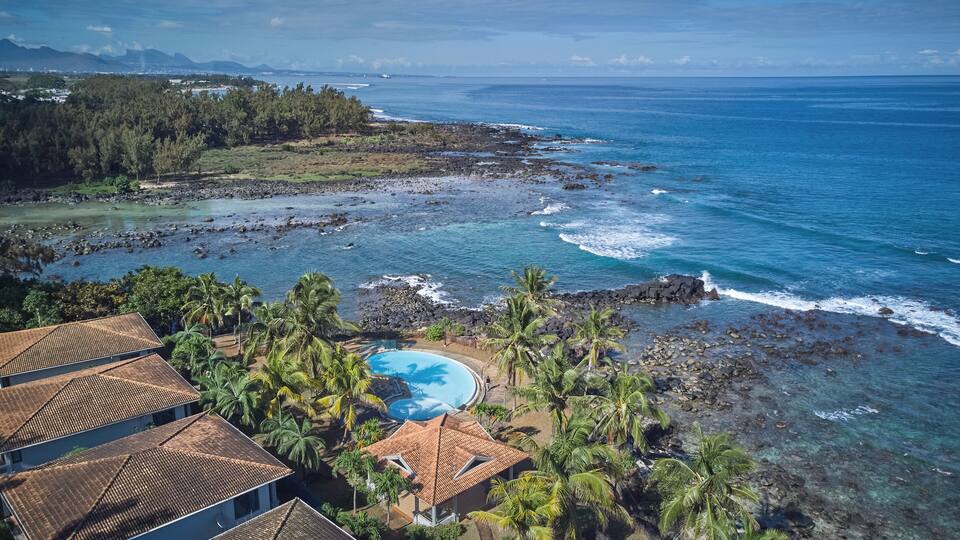 A coastal resort with a clear blue pool surrounded by palm trees and rocky shoreline.