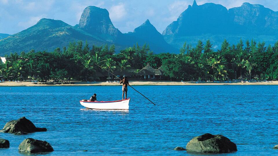 A person in a small boat on a calm sea with a backdrop of mountains and a beach with palm trees.