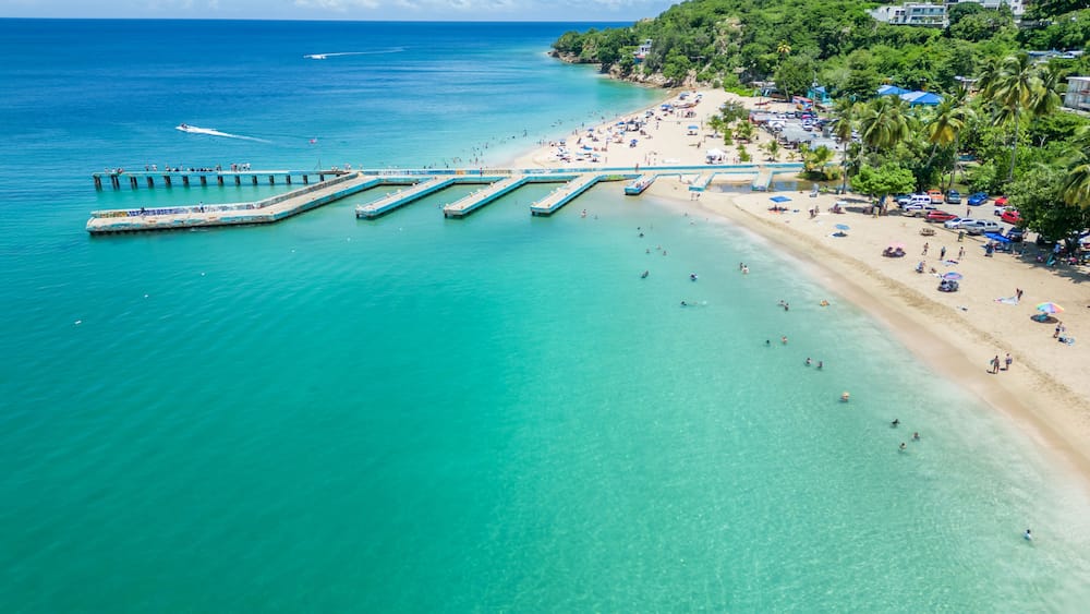 Uma praia com águas turquesa cristalinas, um cais que se estende até o mar, e um morro com edifícios e palmeiras.