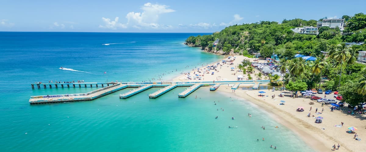 A beach with clear turquoise waters, a pier extending into the sea, and a hillside with buildings and palm trees.