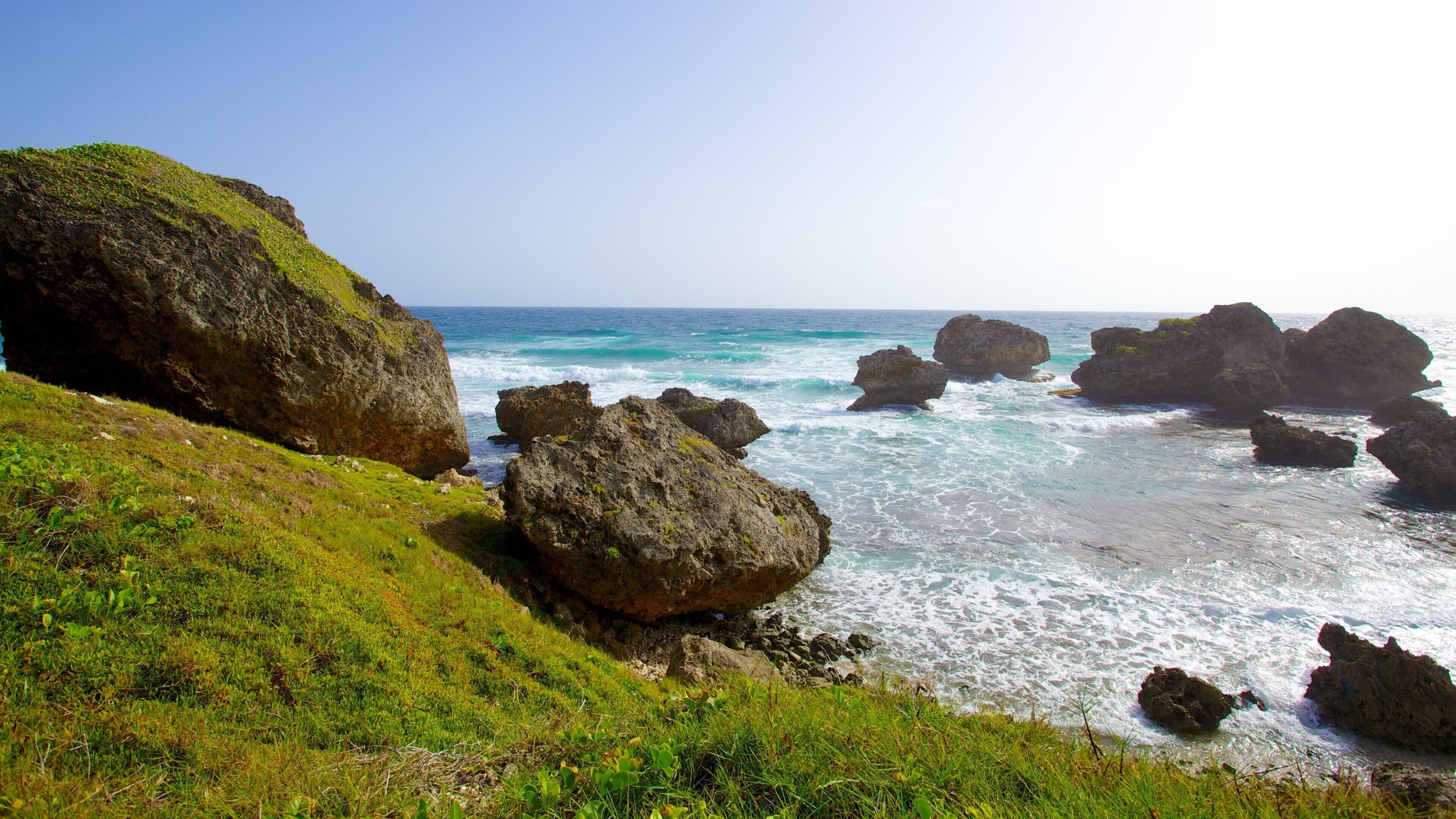 A rocky coastline with waves crashing against large boulders and a grassy hill in the foreground.