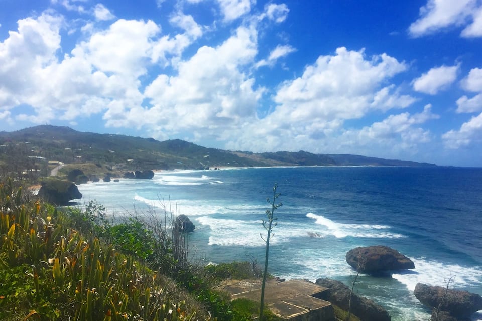A coastal landscape with rocky shores, waves crashing against the rocks, and a clear blue sky with scattered clouds.