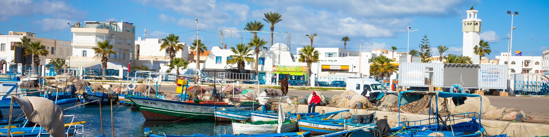 Un puerto con numerosos barcos pequeños, un faro y palmeras.