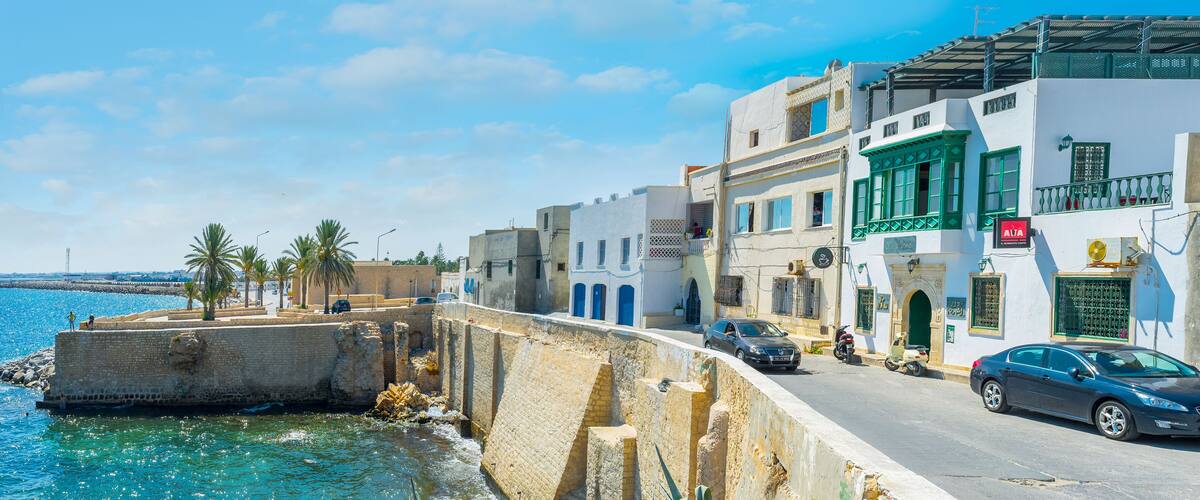 A coastal street with buildings, a sea wall, and parked cars.