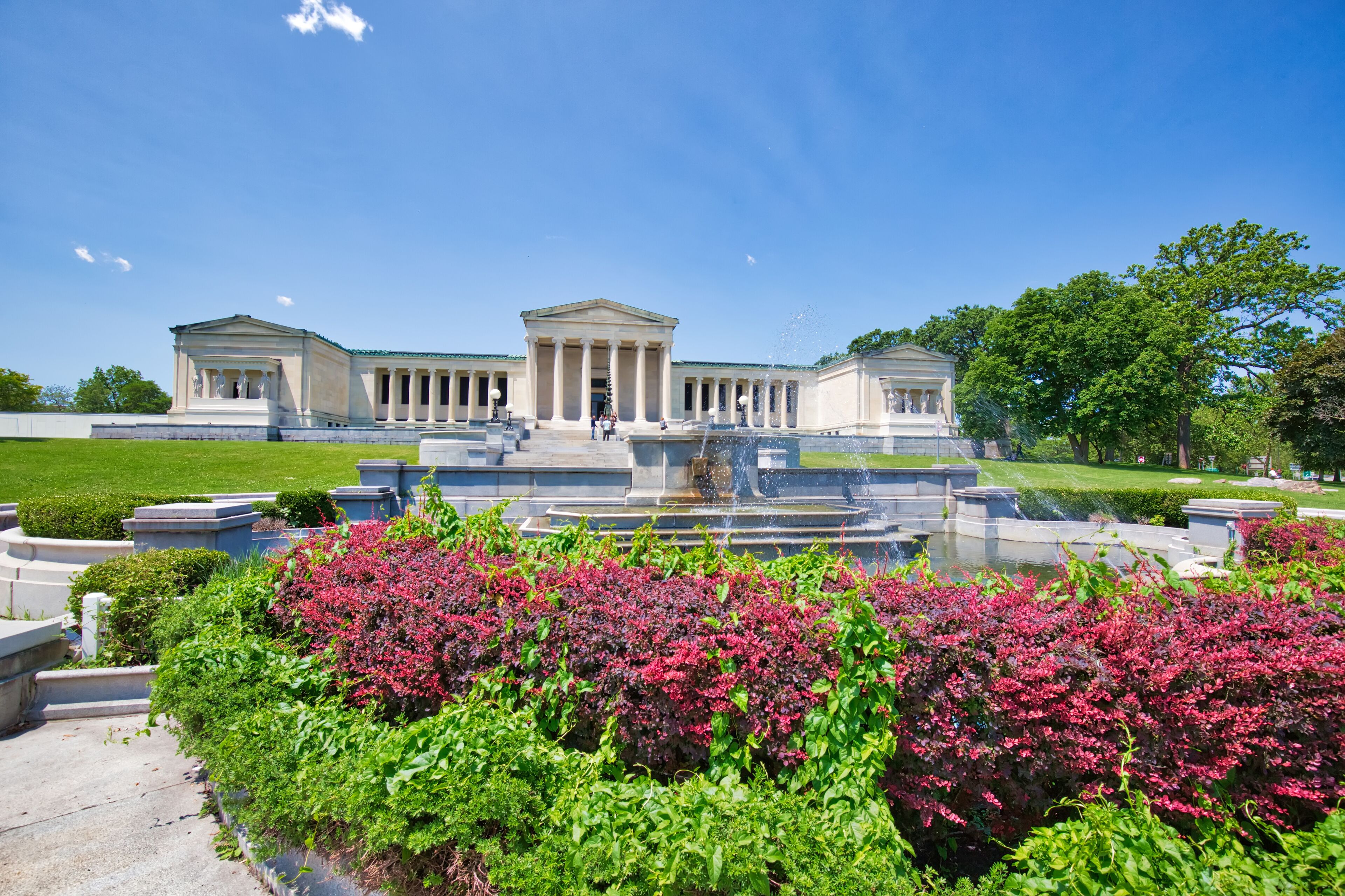 A neoclassical building with a central pediment and columns, surrounded by a fountain and lush greenery.