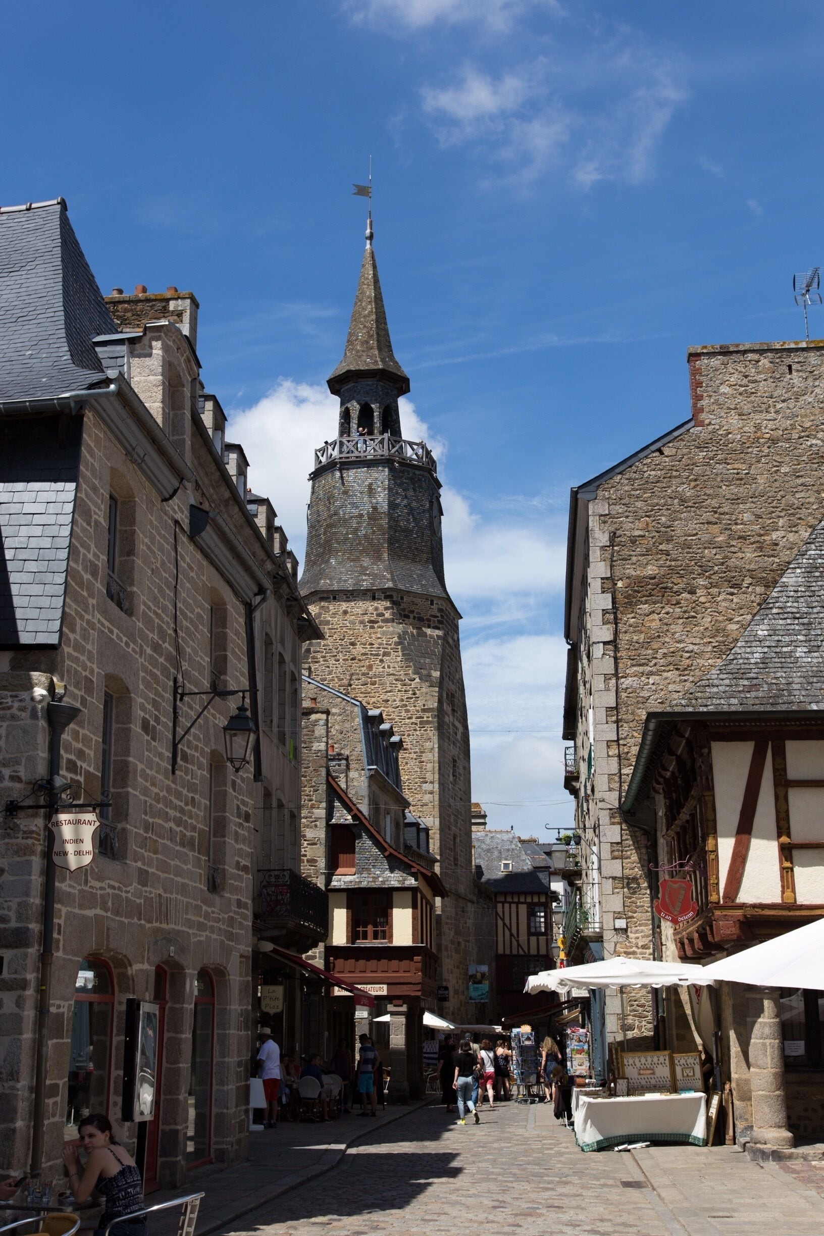 A narrow cobblestone street flanked by historic stone buildings with a prominent tower and a church steeple.