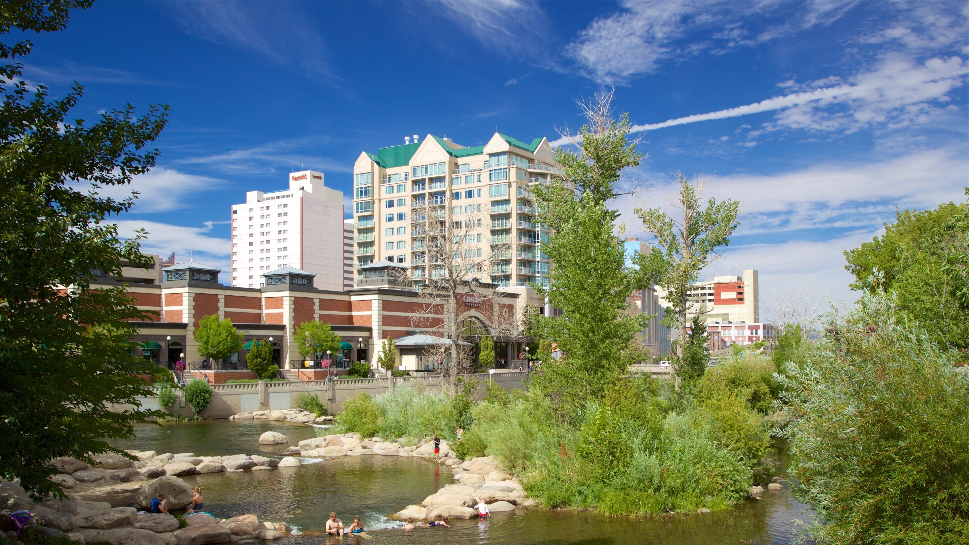 A riverside with rocks and greenery, a hotel building, and a clear blue sky.