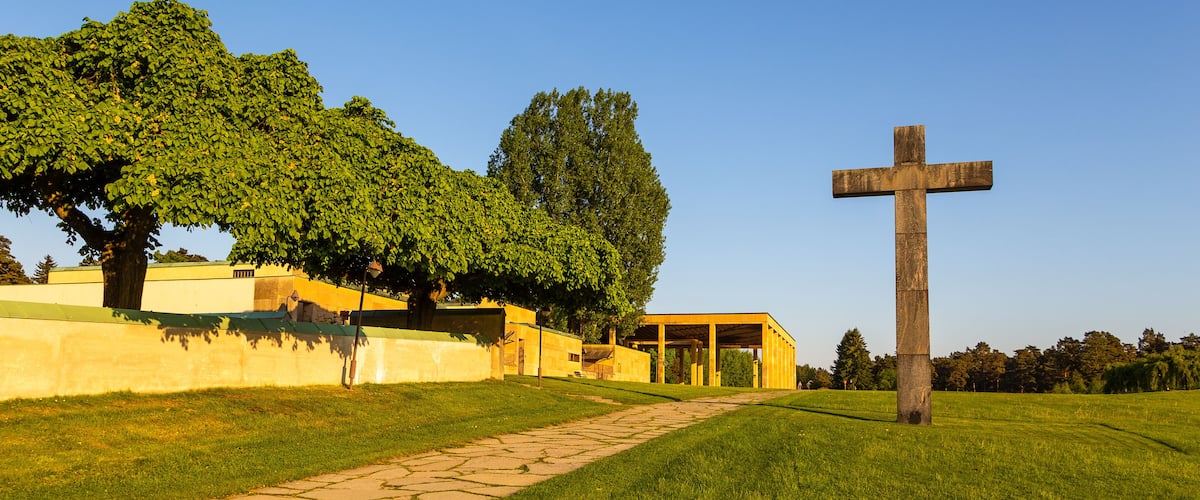 A grassy field with a stone pathway, a wooden cross, and a building with a yellow facade.