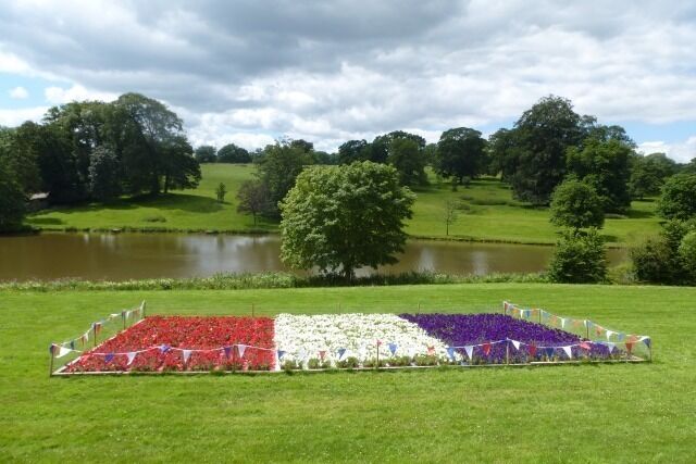 Um campo com uma disposição de flores em formato de bandeira, um lago e árvores ao fundo.