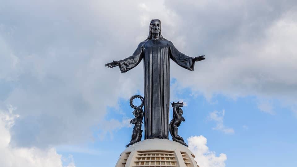A statue with outstretched arms and two smaller figures on either side, set against a cloudy sky.
