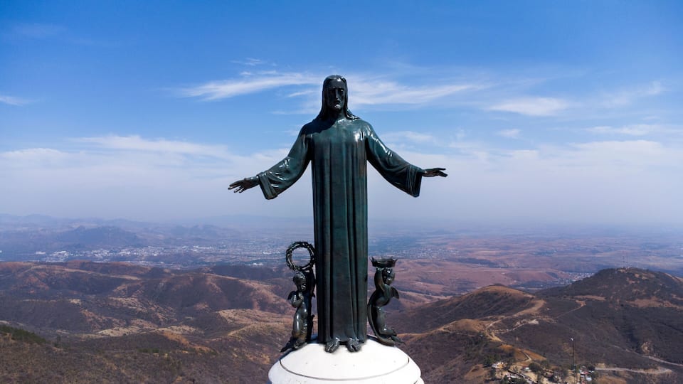 A statue of a robed figure with outstretched arms, flanked by two smaller figures, set against a mountainous backdrop.